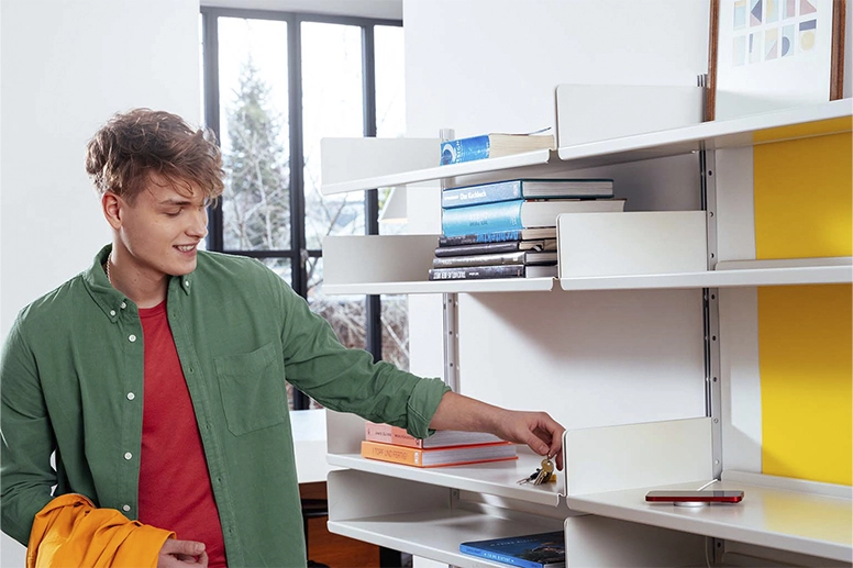 A young man stands in front of a shelf on which a mobile phone lies while being charged with a MagSafe charger.