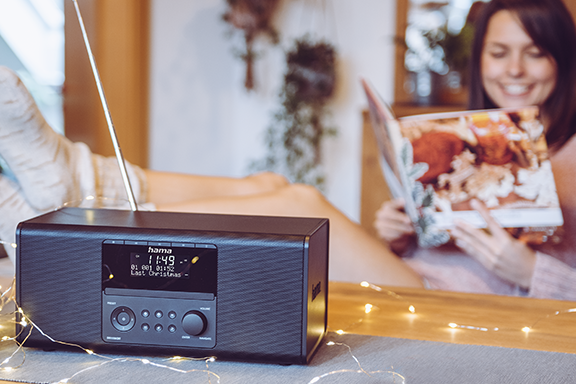 A young woman sits relaxed and in a good mood at the breakfast table, leafing through a magazine, while the Hama digital radio "DR1550CBT" stands on the table and plays music